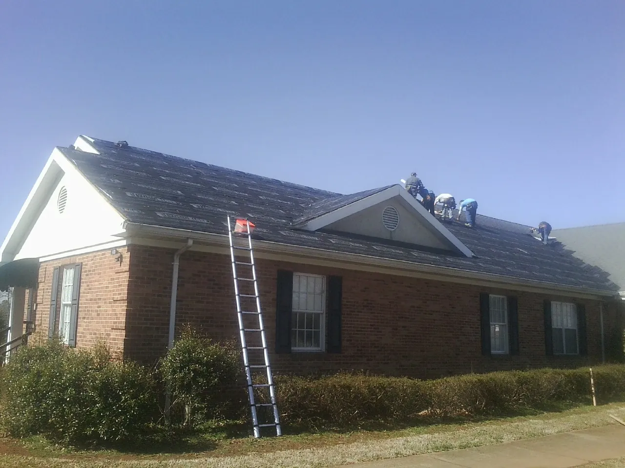 Crew tearing shingles off a brick ranch roof with a ladder set — Metal Roof Repair work by our crew in Falling Waters