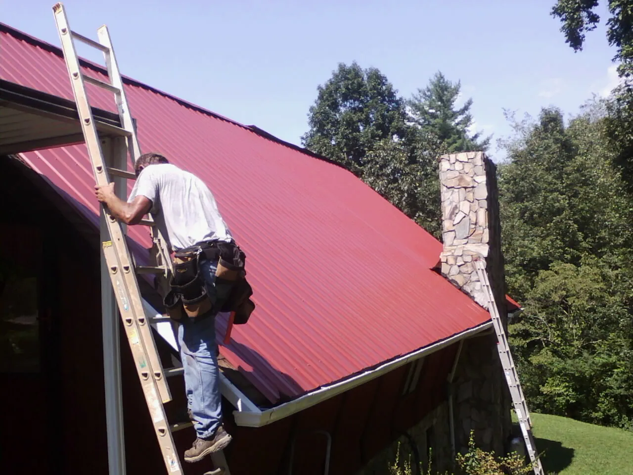 Roofer on a ladder beside a red metal roof and stone chimney — Soffit & Fascia Repair by Roofing Brothers in Falling Waters
